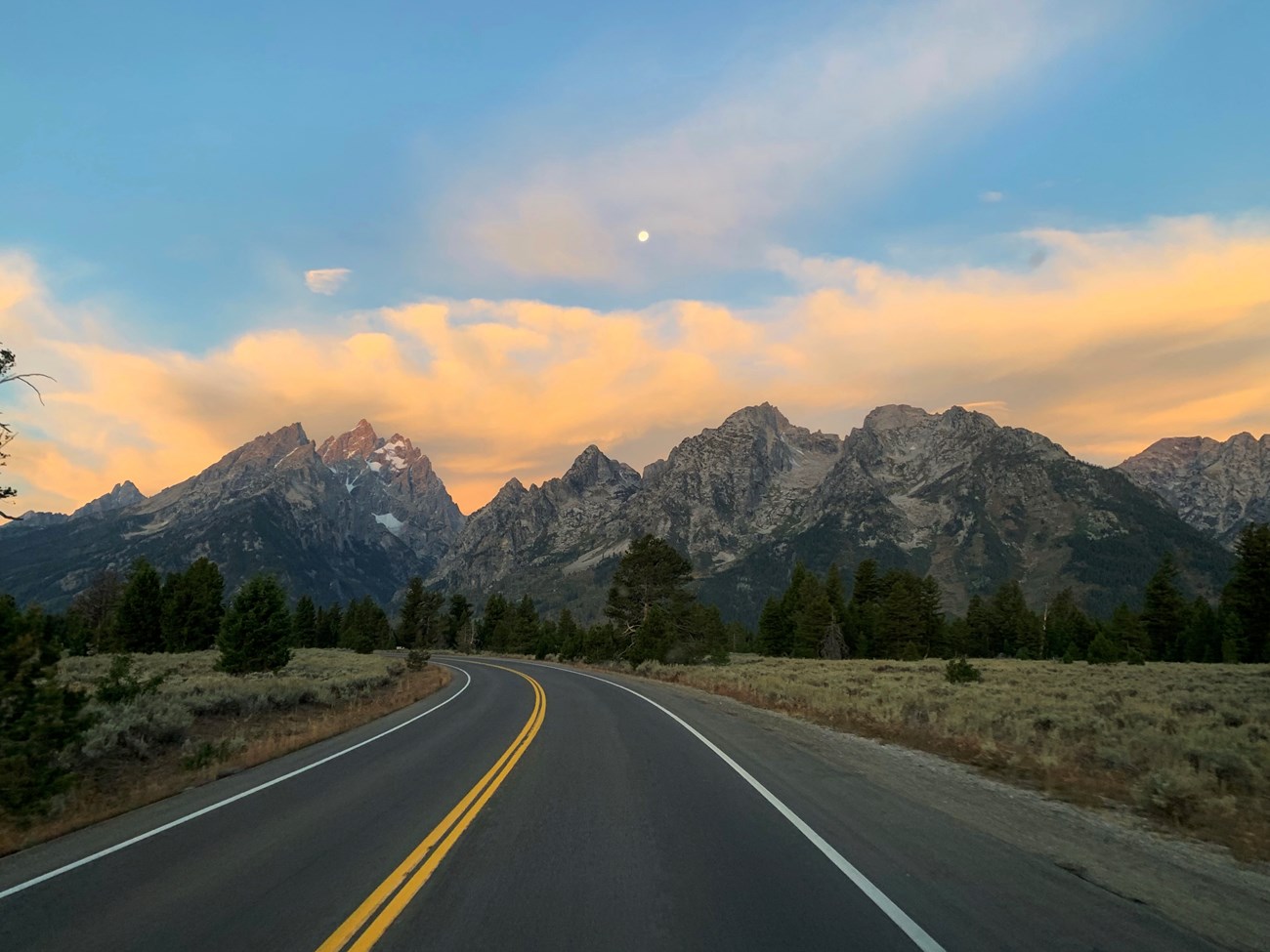 A road headed towards mountains with bright clouds behind them.