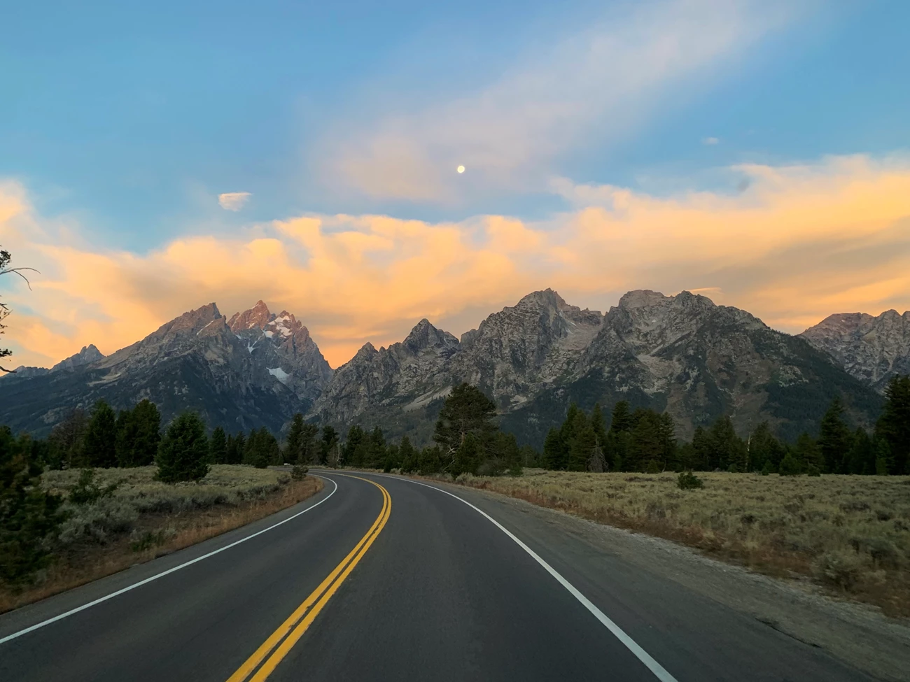 The Teton Park Road A road headed towards mountains with bright clouds behind them.