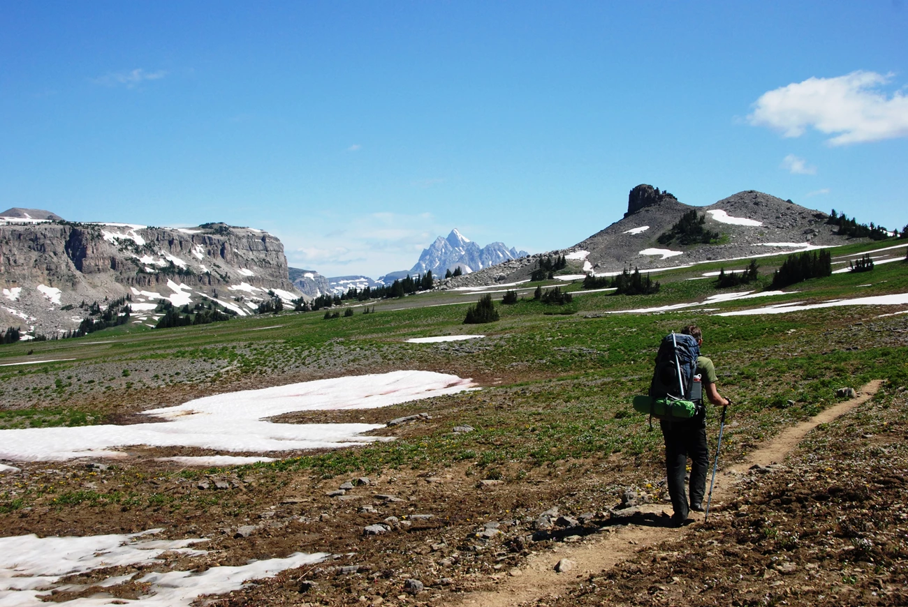 Hiking A hiker with a large backpack walks along a grassy trail towards mountains.