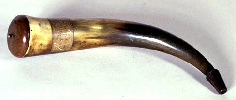 A cow horn modified to hold black powder. There is a label on the horn and a wooden buttplate and stopper at either ends. The horn measures 36 cm long
