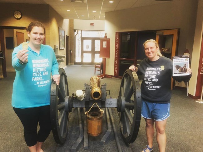 Two women stand with a cannon between them. Both hold Junior Ranger badges