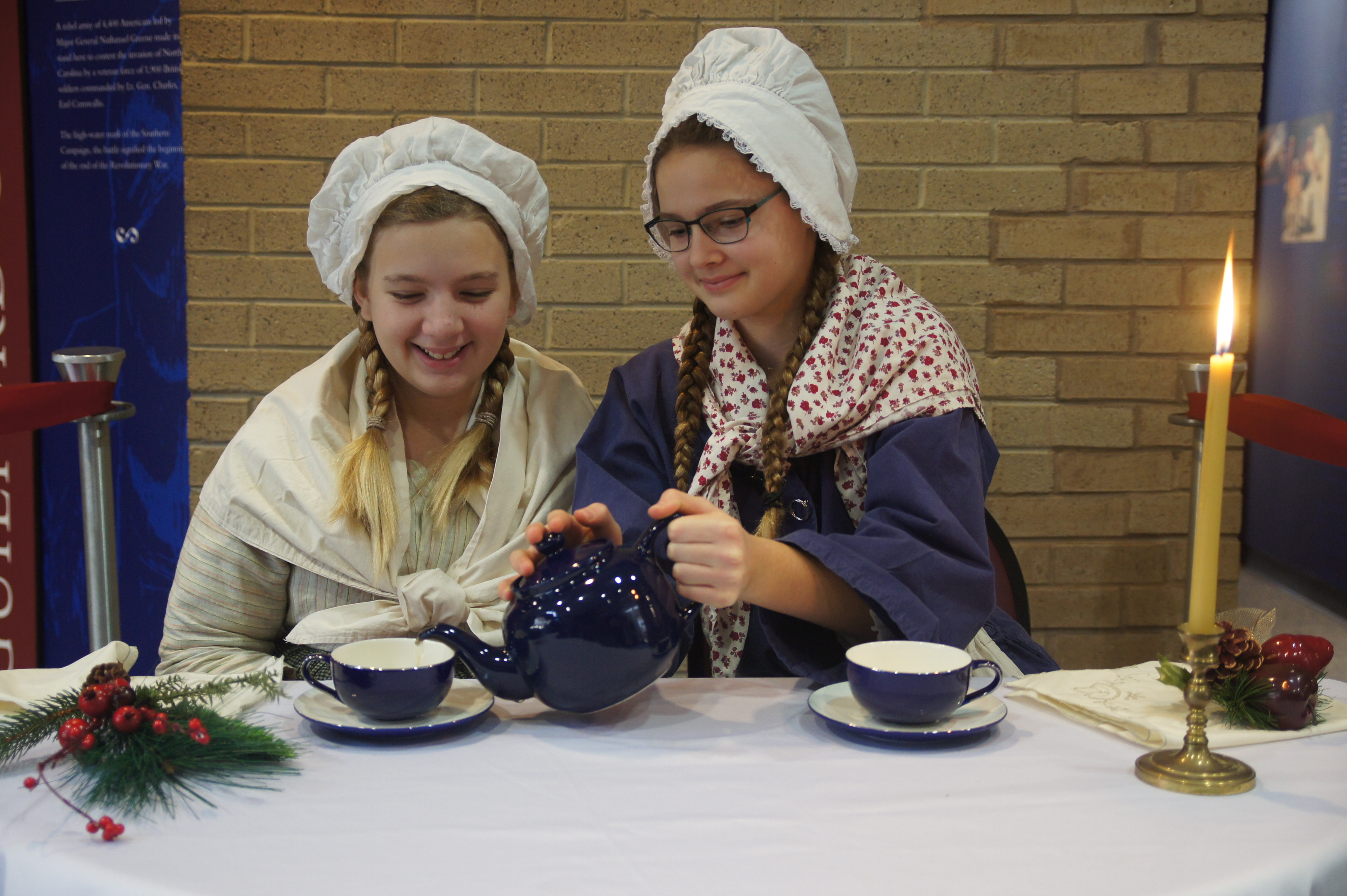 Two girls in colonial clothing pour tea out of a blue teapot