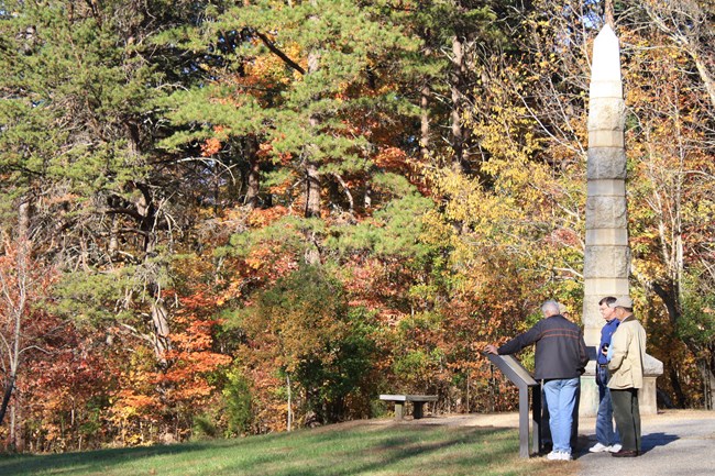 Three men read an exhibit panel and an obelisk monument stands behind them with a forest.