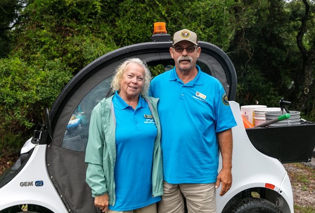 Two campground hosts stand in front of their golf cart and smile at the camera.