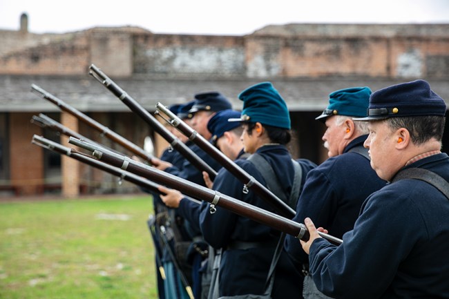 Volunteers and staff members dressed in historic Civil War era uniforms prepare to fire their muskets.