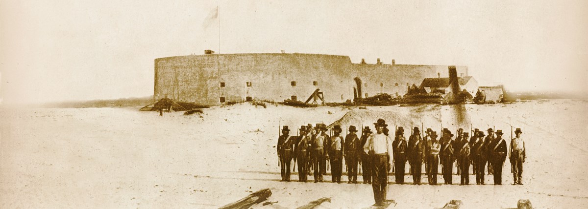 Historic image of soldiers standing in front of a masonry fort. Historic image of soldiers standing in front of a masonry fort.
