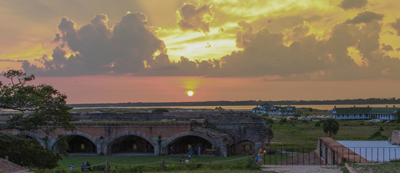 The sun sets behind an island scene with a masonry fort in the foreground.