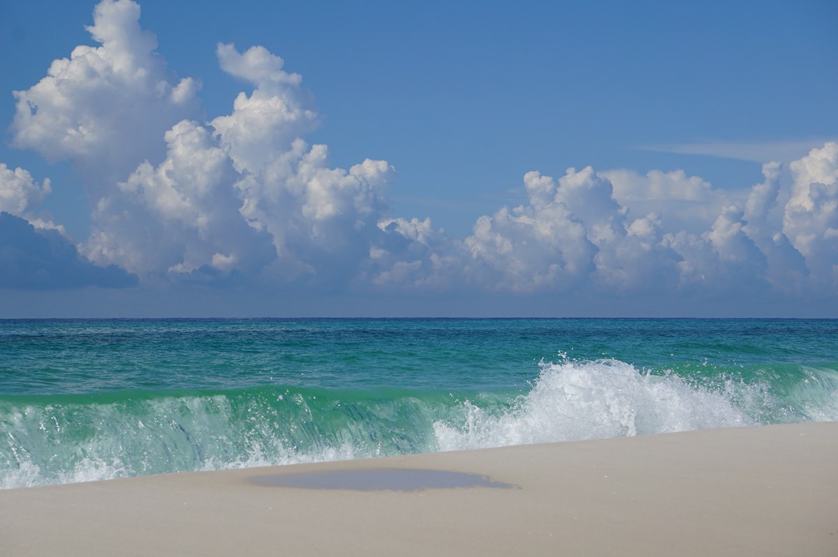 Turquoise waves crash onto a white sand beach. Blue sky and white clouds in the background.