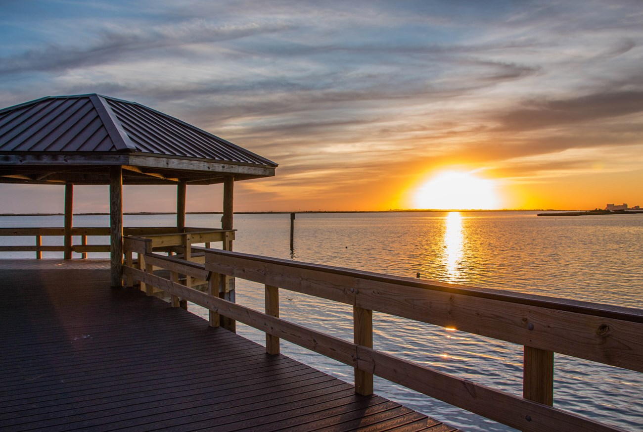 A brown fishing pier stretches over reflective water. A yellow sun sets in the blue evening sky.