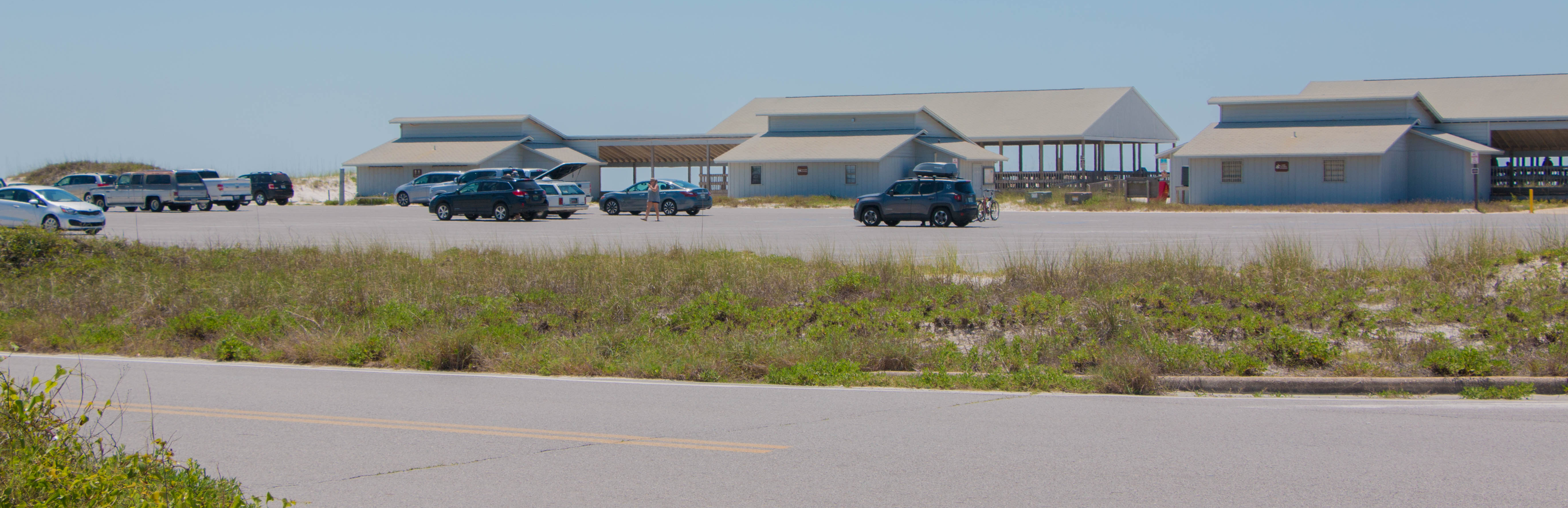 Several one story buildings and pavilions stand on the edge of a beach behind a parking lot. Several one story buildings and pavilions stand on the edge of a beach behind a parking lot.