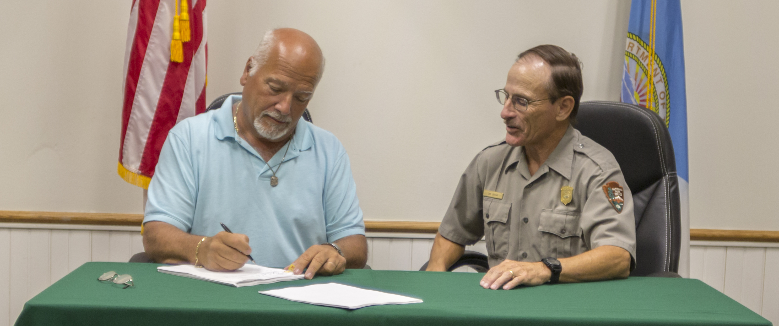 061318_ContractSigning-BrentEveritt_03 Two men sit a table, one signs documents as the other looks on.