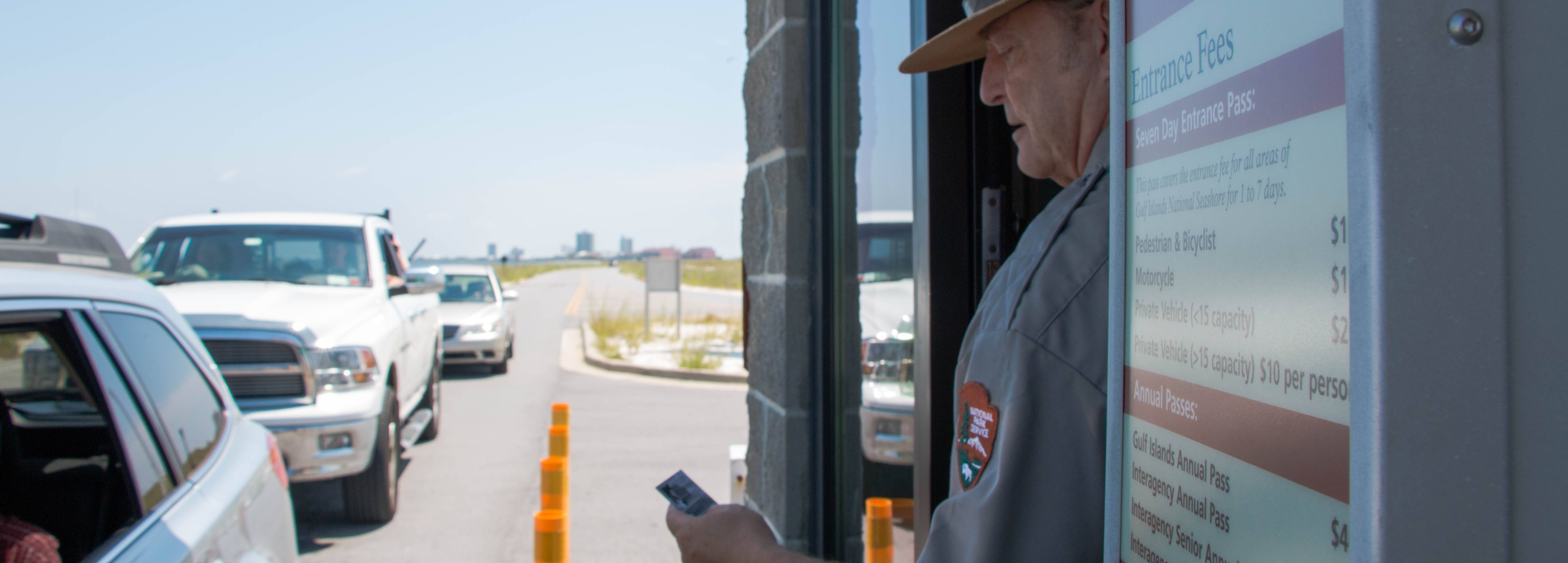 A park ranger checks a pass at the entrance of a park, a line of cars extends into the distance beyond the station. A park ranger checks a pass at the entrance of a park, a line of cars extends into the distance beyond the station.