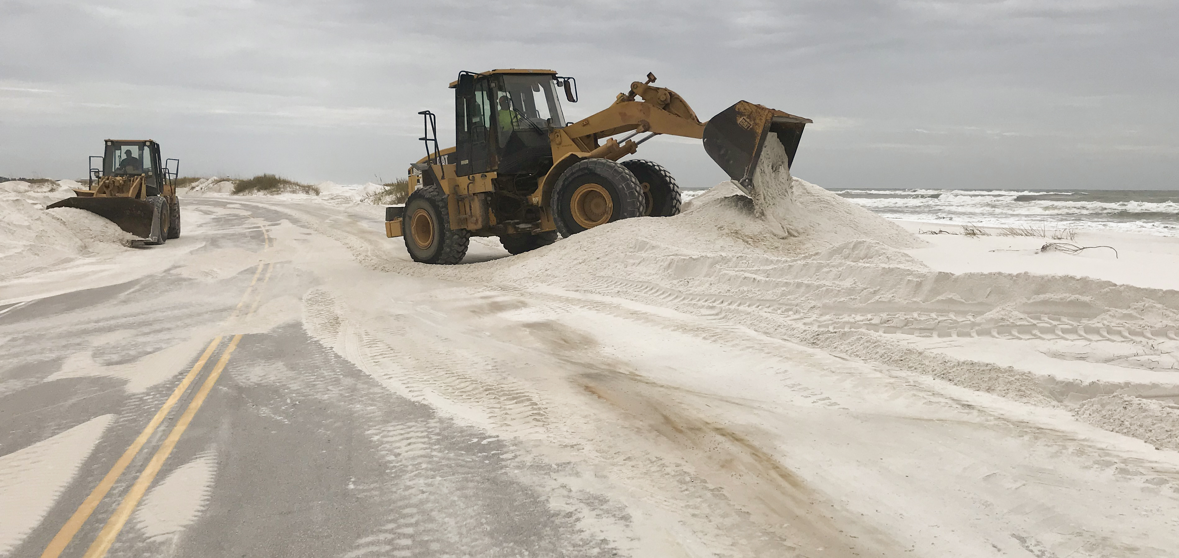 Two heavy machines move sand off of a buried two lane road. Two heavy machines move sand off of a buried two lane road.