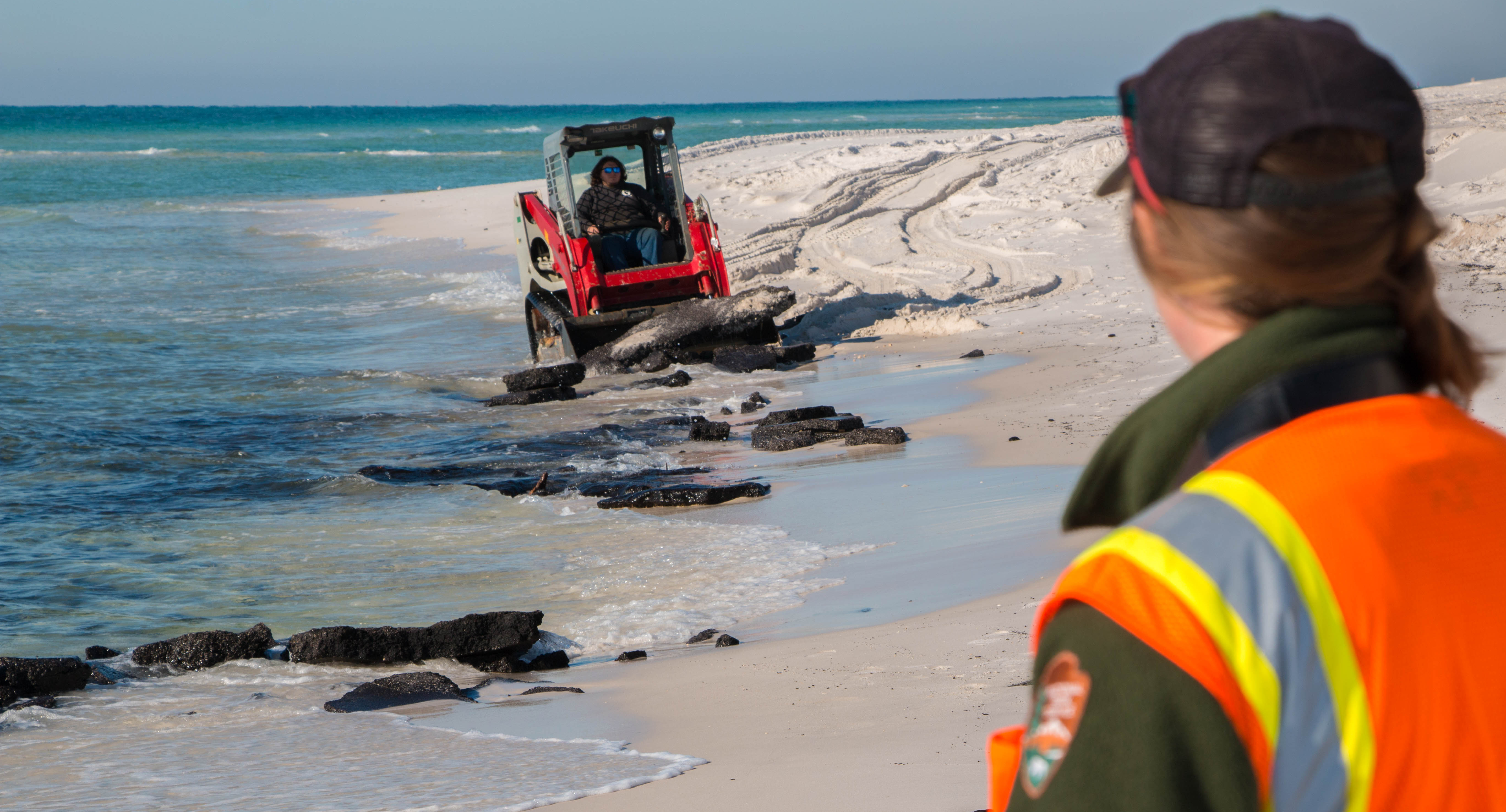 A small front-end loader picks up asphalt debris from the beach at the water line as a ranger looks on. A small front-end loader picks up asphalt debris from the beach at the water line as a ranger looks on.