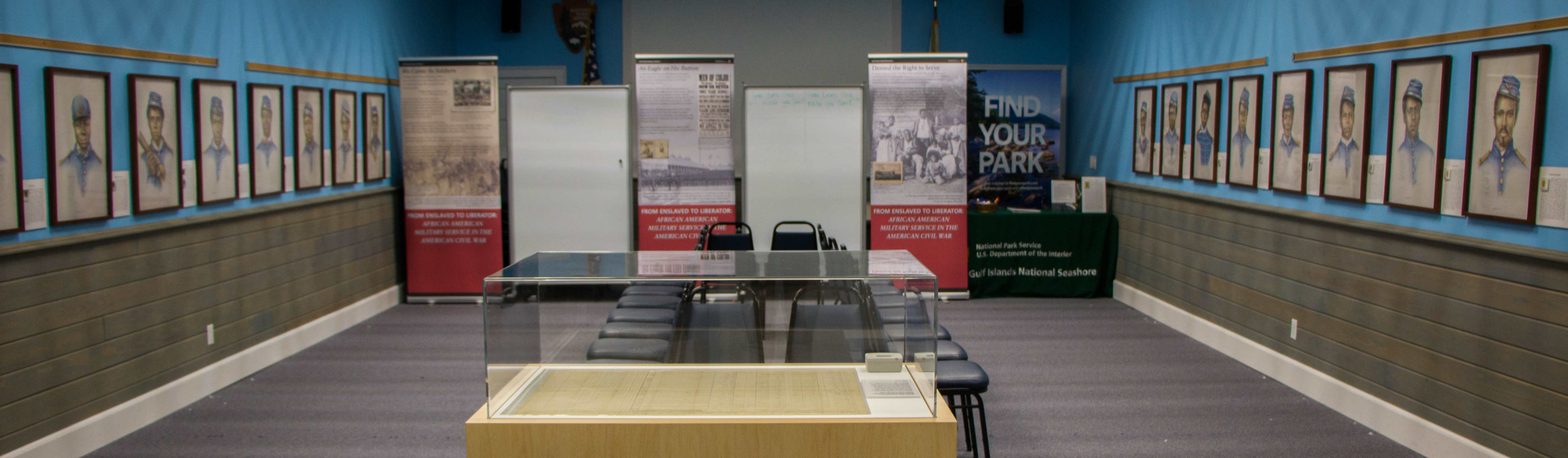 Hand drawn portraits line a blue room on either side, at the end of the room informational banners stand upright, and in the foreground an exhibit case holds a historic document.