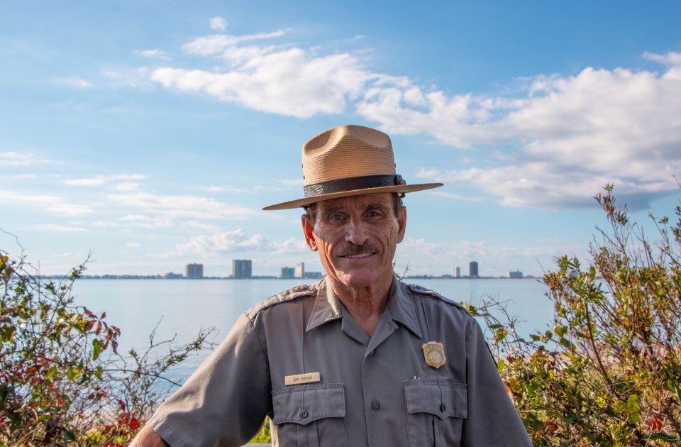 A man in a grey National Park Service uniform shirt with a tan hat standing in front of blue water and sky.