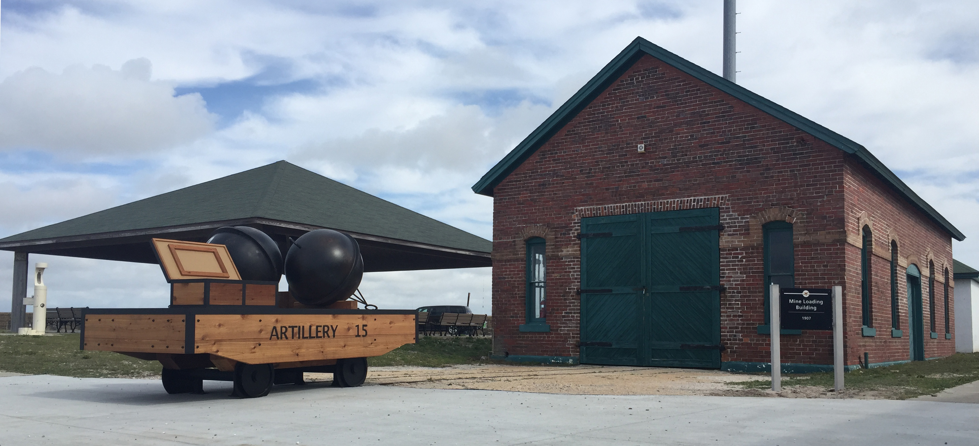 A reproduction rail-cart stands in front of a one story brick building, a sign to the right identifies it as the Mine Loading Building, a shade shelter can be seen in the distance.