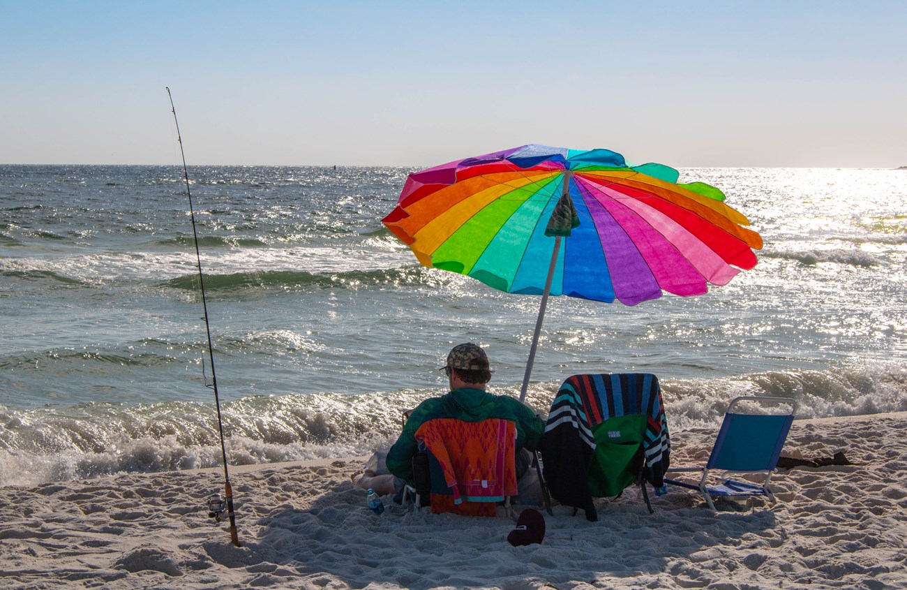 A man sits under a rainbow umbrella with a fishing pole in the sand to his left and the ocean in the background.
