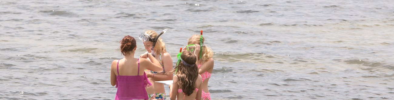 Four women adjust snorkeling gear while standing in water.