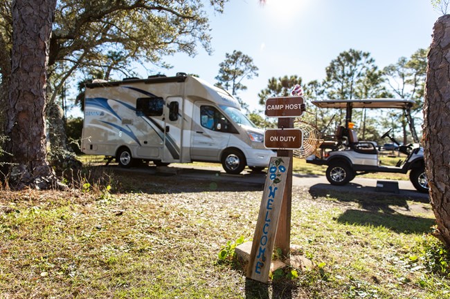 An RV stands behind a Campground Host sign lit by the sun.