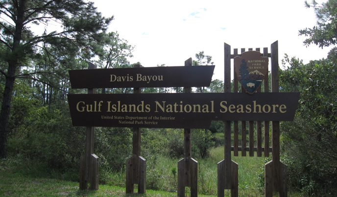A National Park Service sign for Davis Bayou stands in front of a maritime forest. A National Park Service sign for Davis Bayou stands in front of a maritime forest.