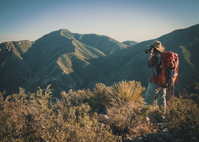 A woman wearing a bright backpack holds a camera while looking down into a wide desert mountain canyon