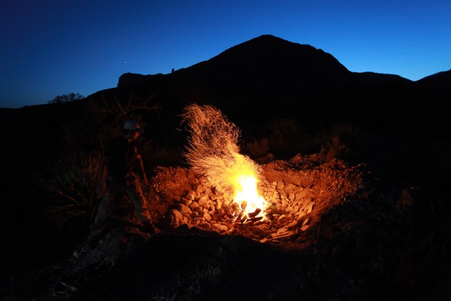 A nighttime photograph of a man standing next to a fire in a shallow pit