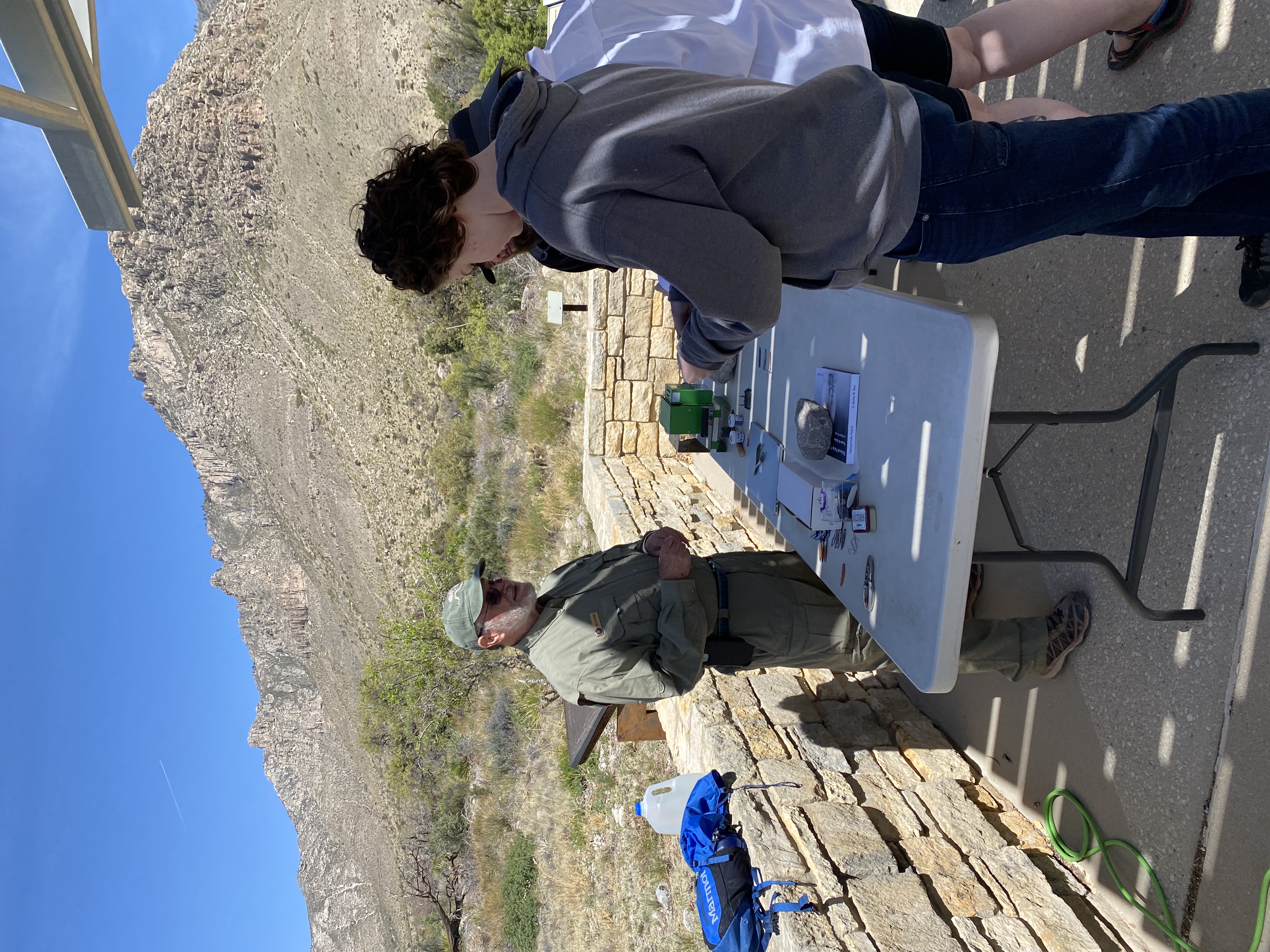 Two people standing at a desk with a man dressed in a brown uniform against large mountains.