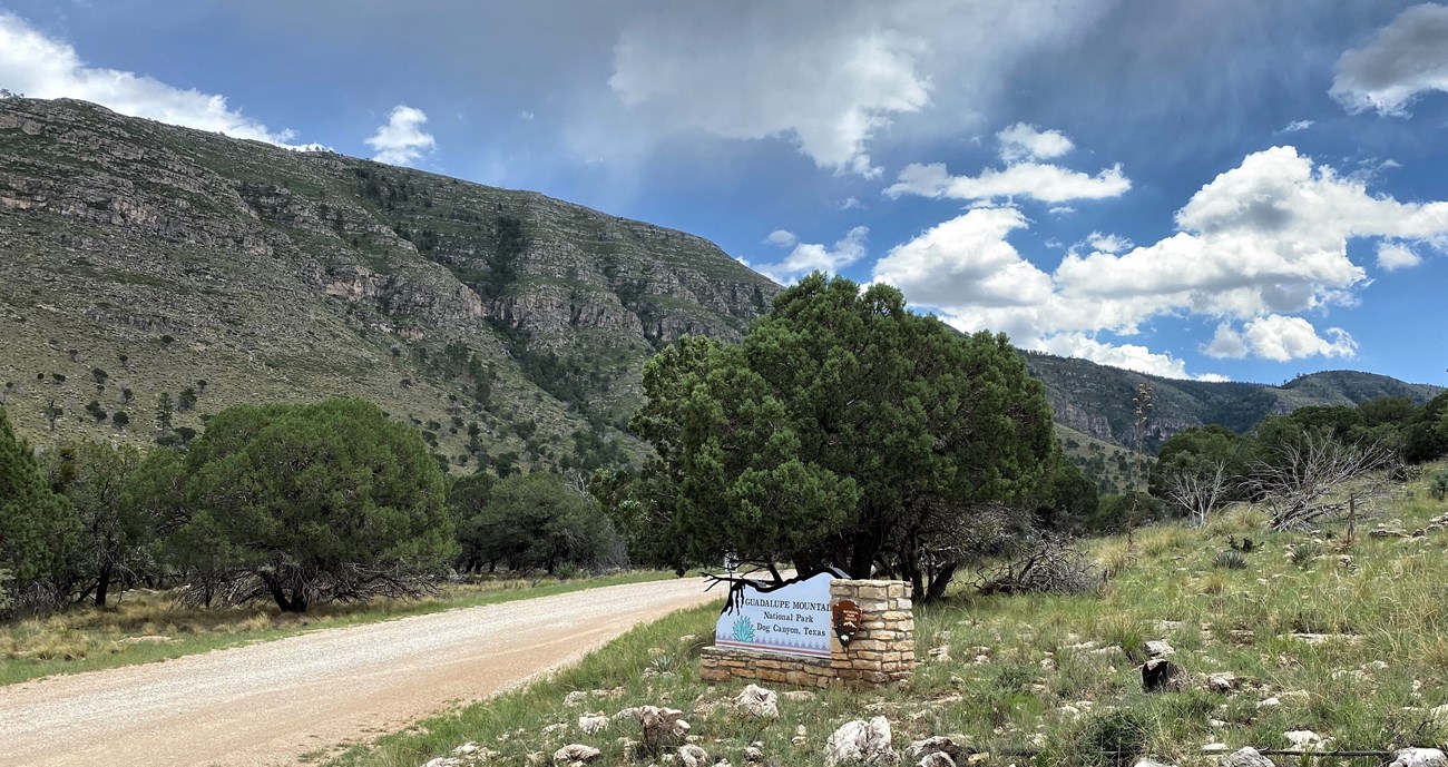 A dirt road leads toward a tall desert canyon with a park entrance sign nearby