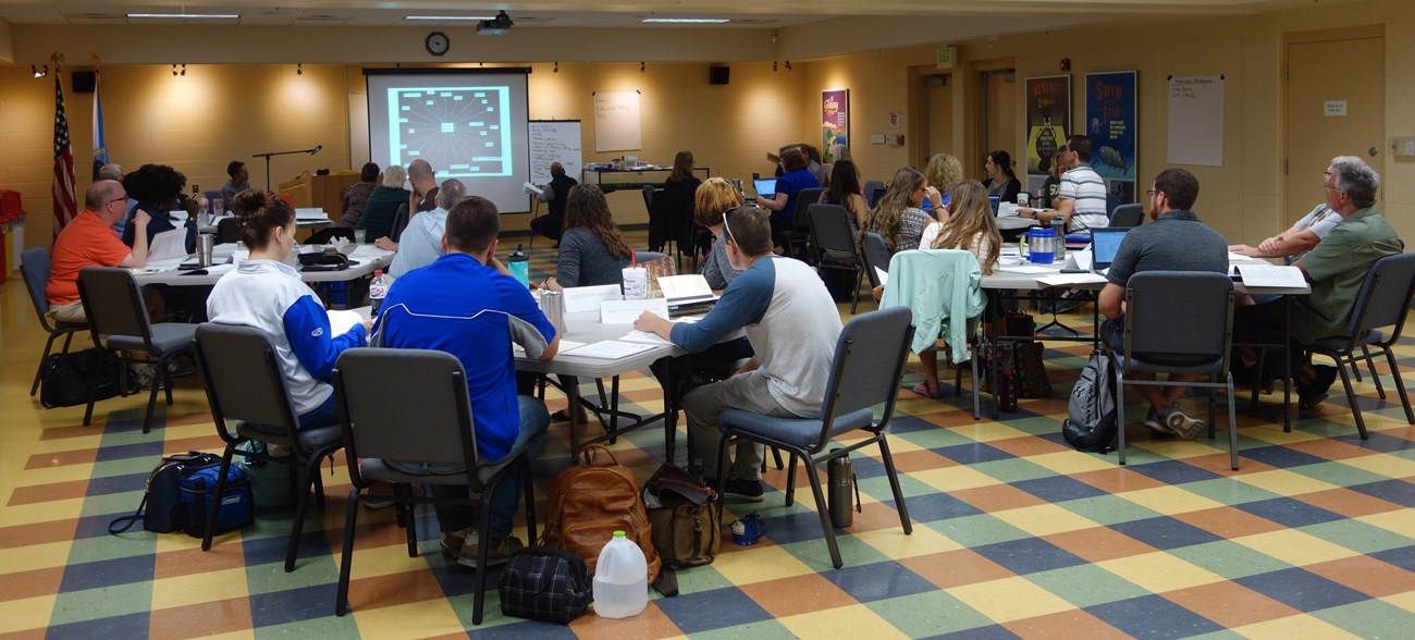 People (teachers) sitting around multi tables watching a presentation.
