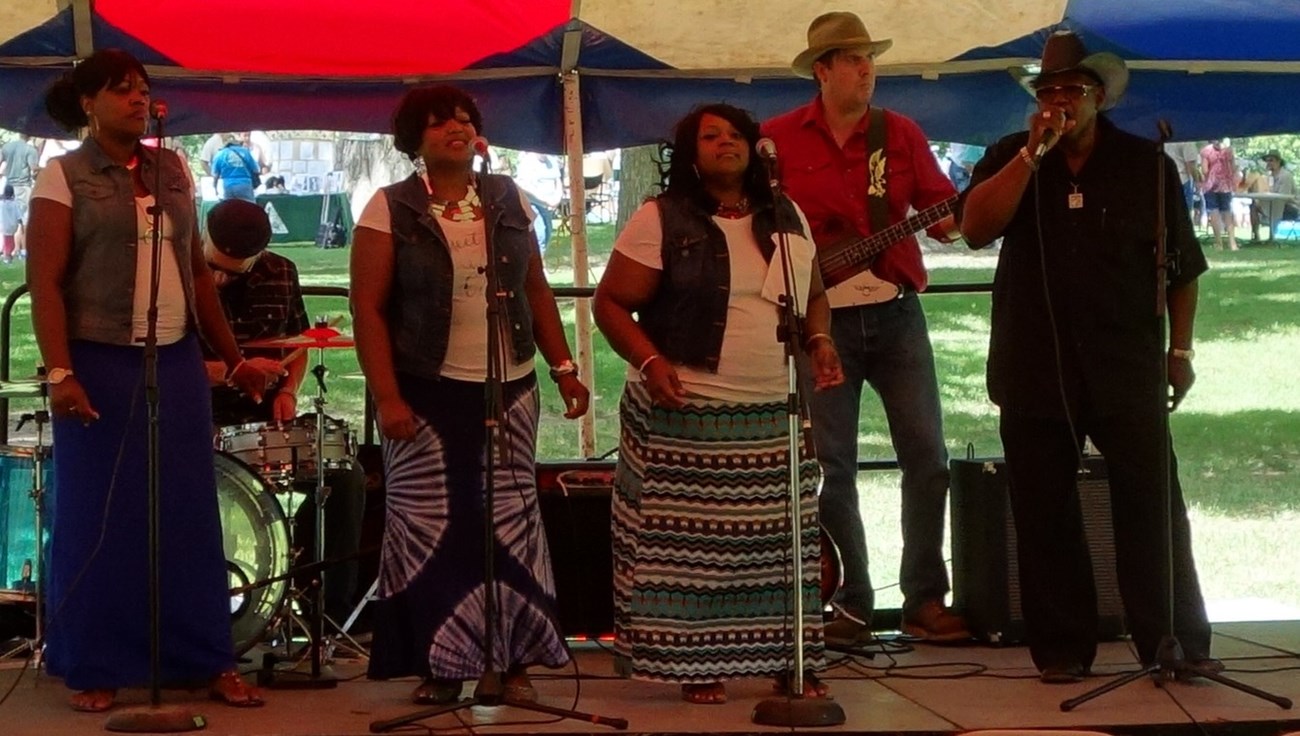 Band members on an outdoors stage. Three women singing with microphones. A man wearing a bat cowboy's hat and signing with a microphone.  A man wearing a red shirt playing a guitar. A man playing a keyboard. A man playing a drums.