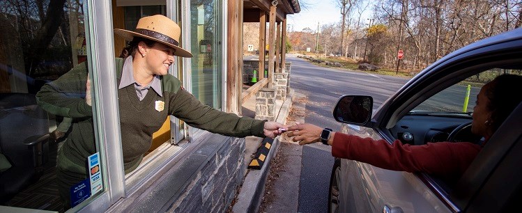 On the left, a park ranger reaches out the window of the fee booth to take a card being handed to her by a visitor, on the right, who is reaching out of the window of her car.