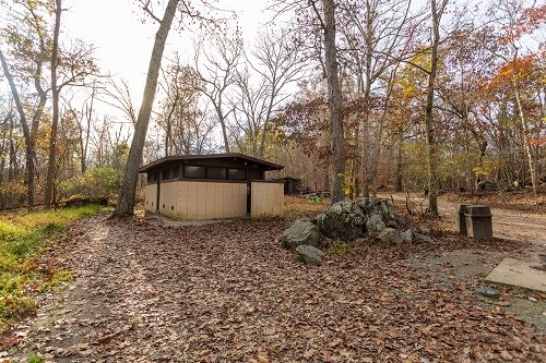 A one-story tall restroom, made from white, painted cinderblocks and brown painted wood, standing on the side of a dirt path, next to a small outcropping of rocks and among many trees with orange and yellow leaves.