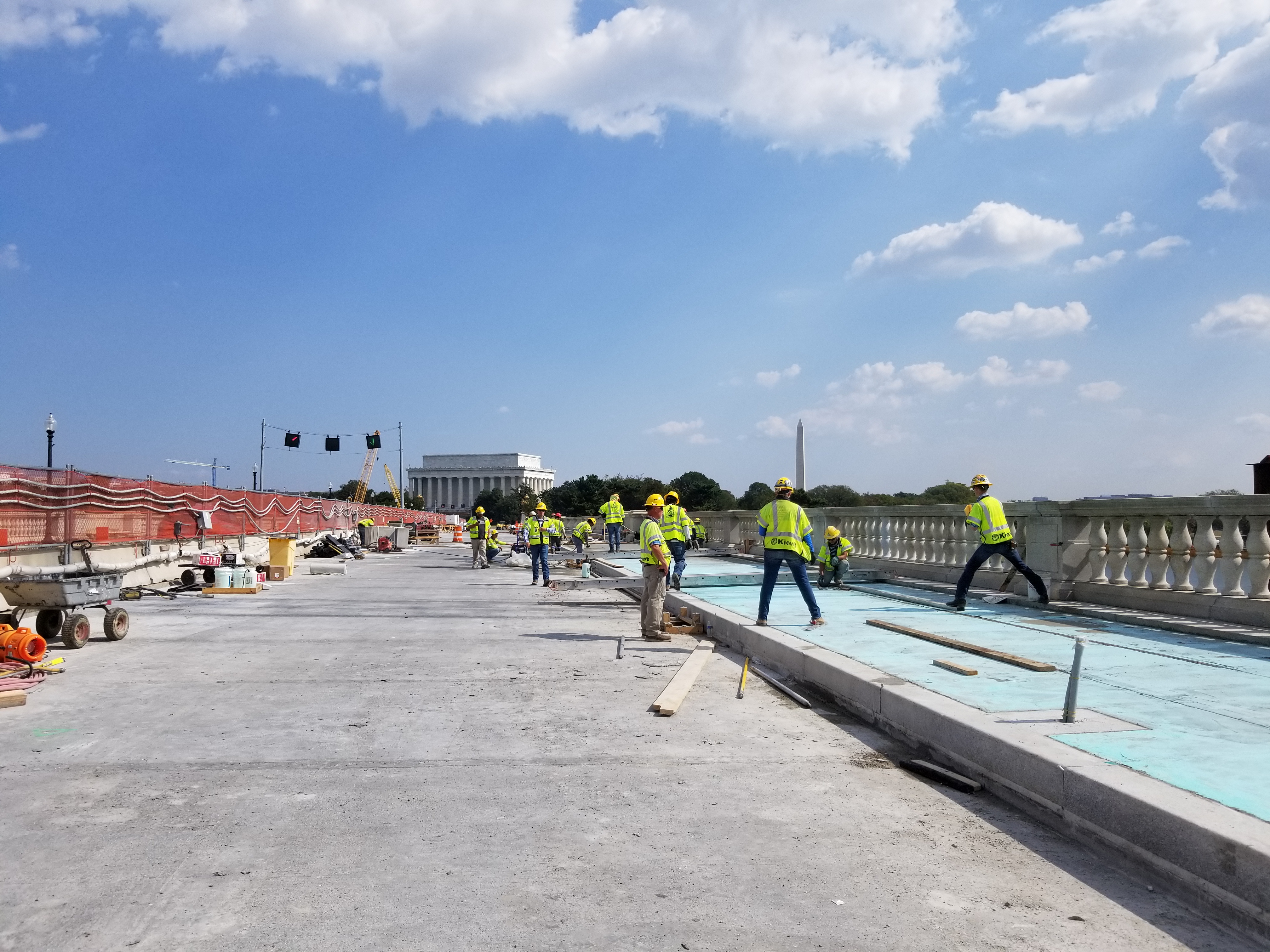 Construction on October 2, 2019 Construction workers prepare sidewalk on Arlington Memorial Bridge with National Mall monuments behind. Bridge deck is exposed concrete with temporary wall on side near active traffic and historic granite balustrade on the other side.