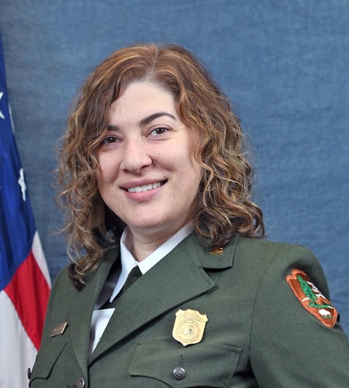 Jennifer Madello A woman with reddish brown wavy hair smiles in a park ranger uniform. A portion of the American flag can be seen on the left side. The background is a solid denim blue.