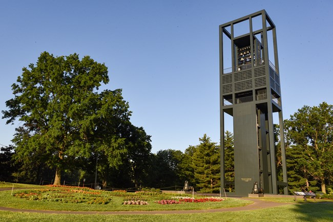 A dark, metal bell tower is surrounded by grass, trees, and a manicured flower garden.