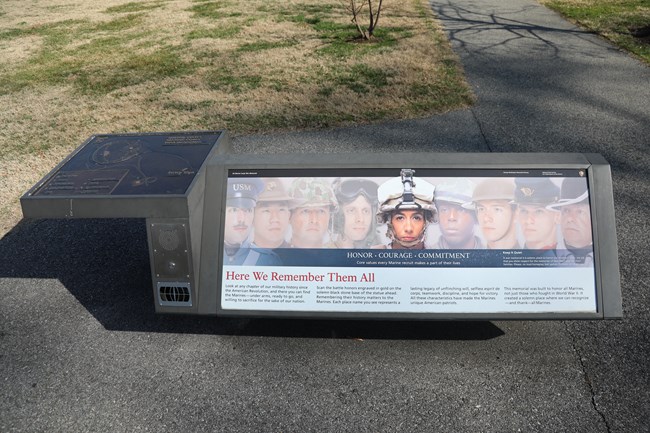 An informational panel on a paved sidewalk with a bronze relief map of the memorial plaza with a button and speaker below.