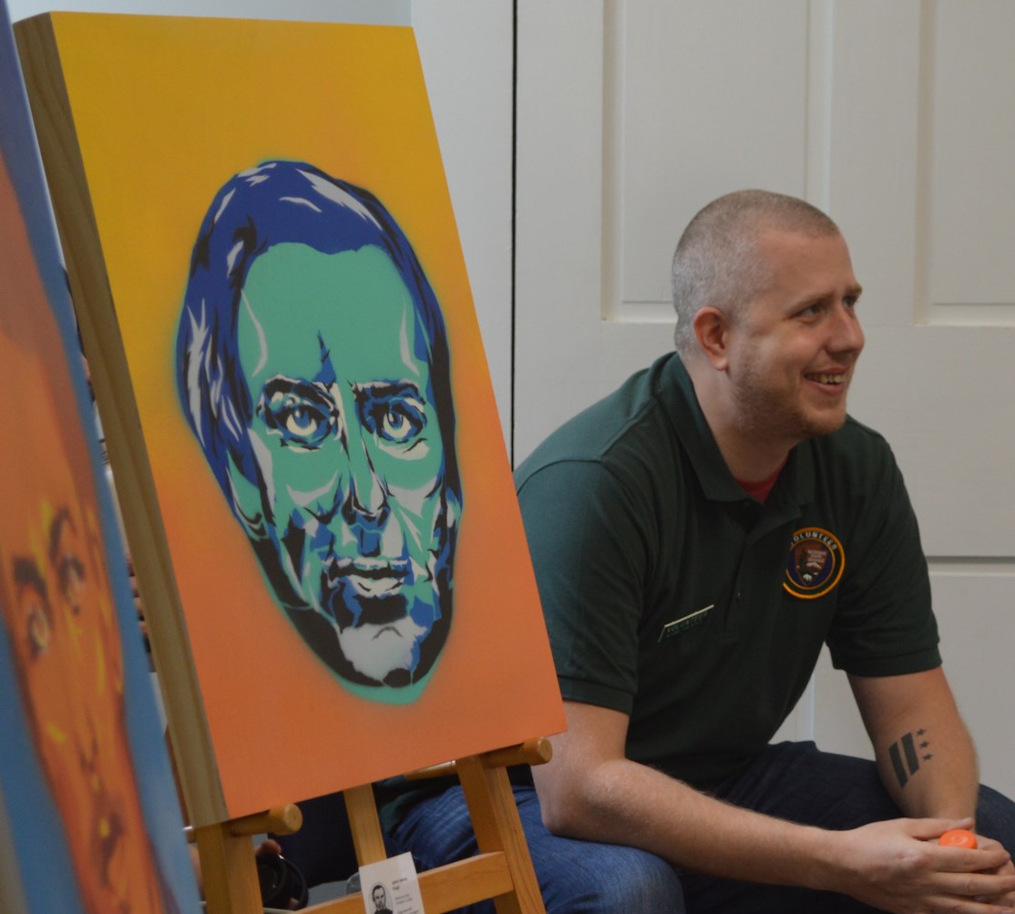 Man in green shirt sits next to portrait painting