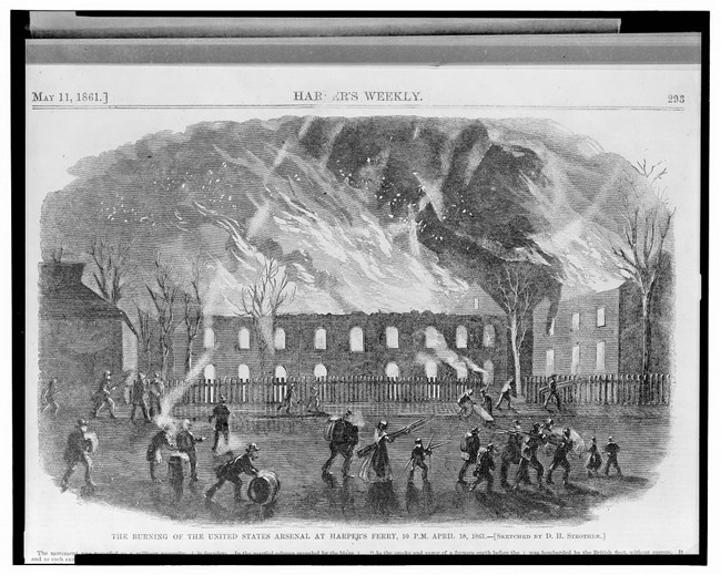 a large building burns in the background as a number of people mill around in front, many with armfulls of weapons and other salvage