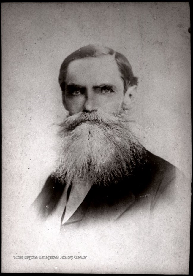 Black and white photograph of a man from the chest up with a very large beard and pointed handlebar mustache.