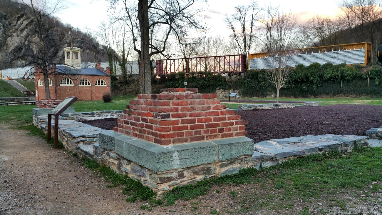 Foreground corner of a brick ruin, background a brick building with cupola and further back a train on an elevated trestle