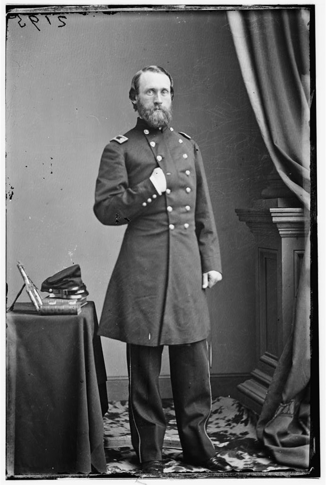 bearded man in dark colored, double breasted military frock coat stands next to a waist height table, one of his hands is tucked into the coat