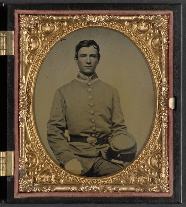 young man in single-breasted military frock coat, the number 2 on his collar, and the letter A on his hat, which is sitting on his lap.