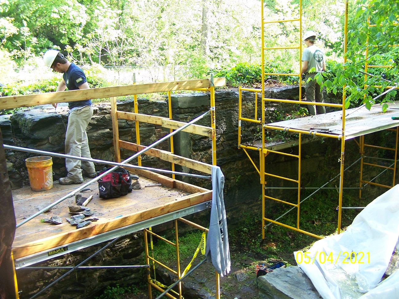 Two workers in hardhats stand on scaffolding to repair stonework.