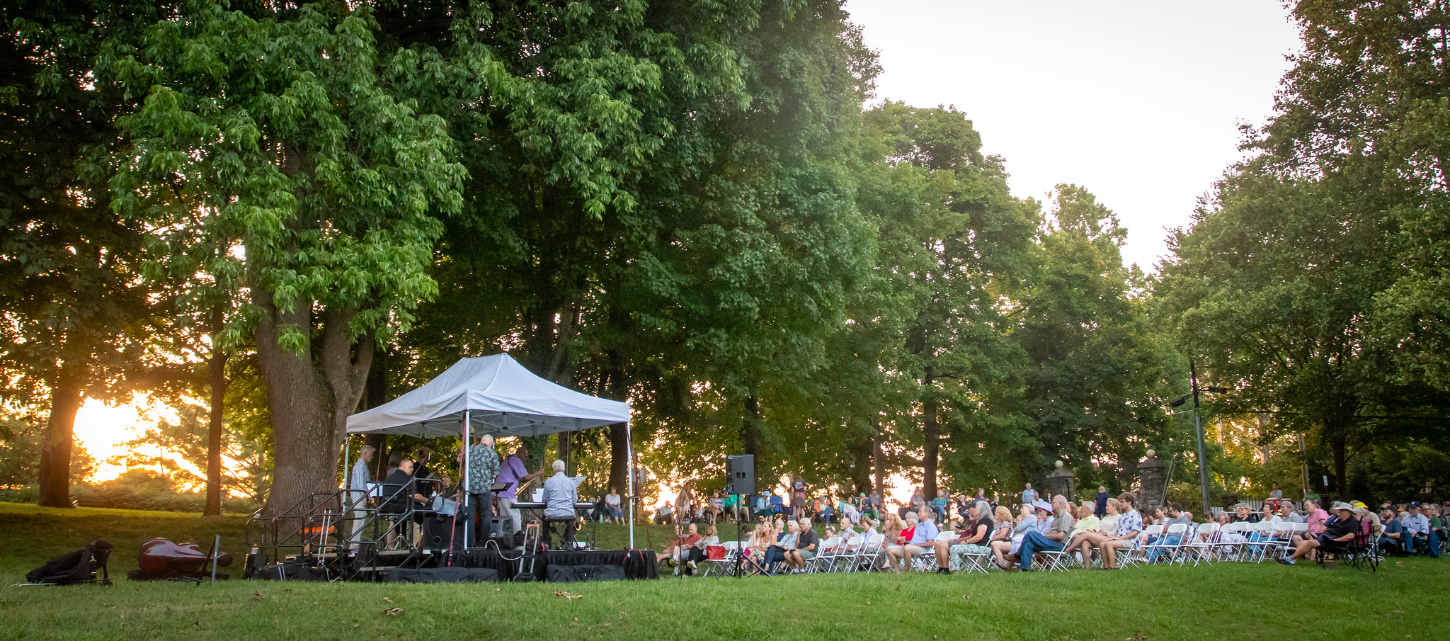 Musicians under a tent on a lawn play to a crowd of people sitting in chairs at sunset.
