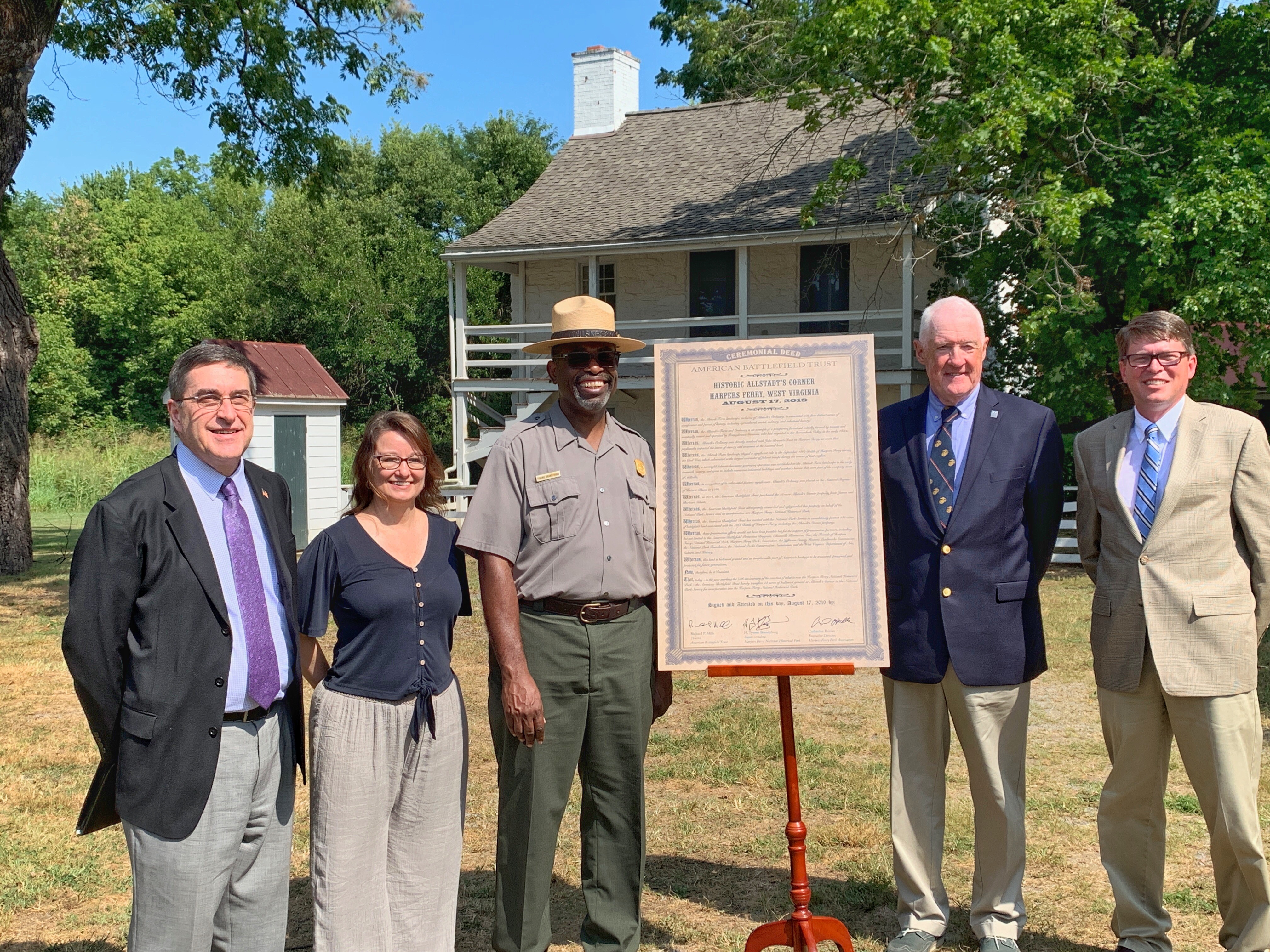 speakers from the dedication ceremony pose with the ceremonial deed five people stand outdoors next to an easel which holds a ceremonial deed; behind them is a historic building
