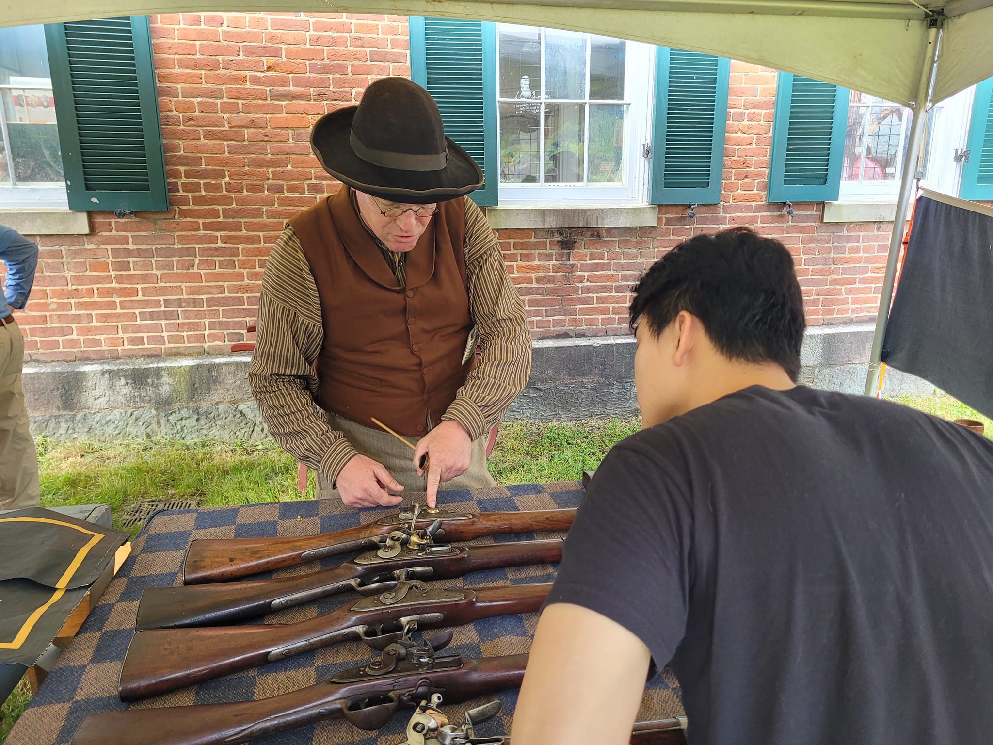 Am man in early 19th century attire shows details of a weapon to a visitor.