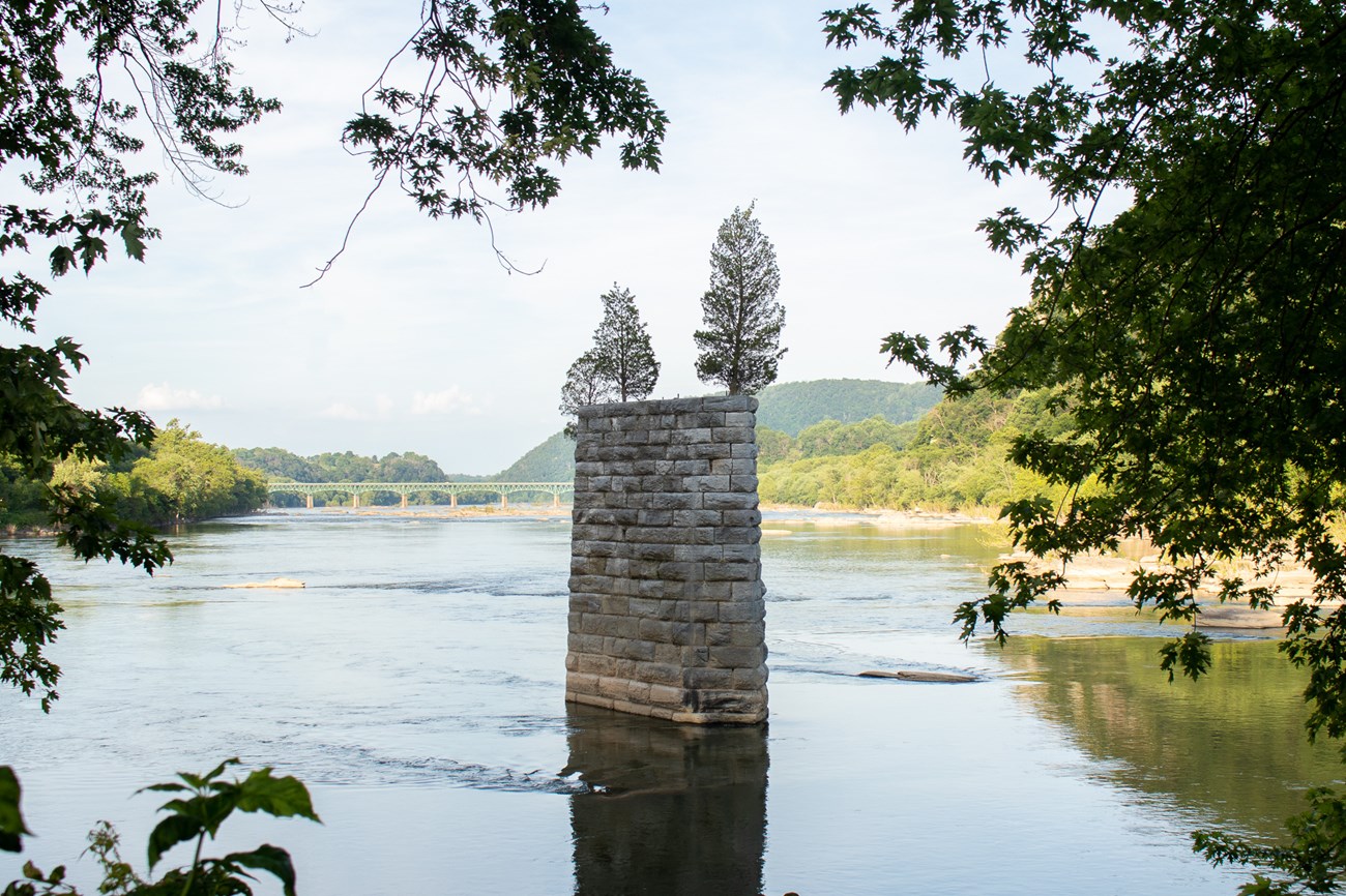 A river flows around a tall bridge ruin made of stone, with trees growing atop it
