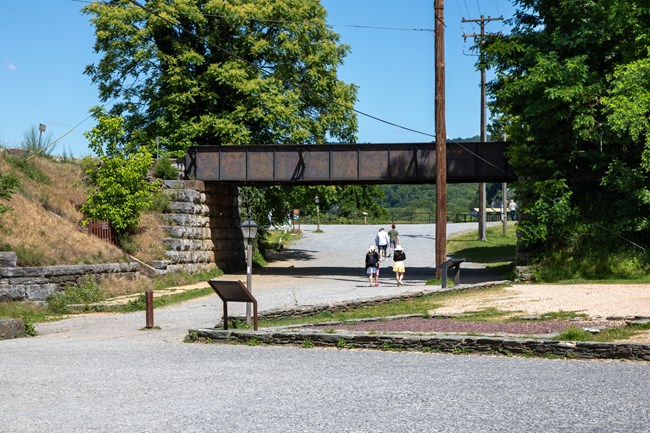 A railroad bridge passes over a wide gravel road with people walking towards a railing with signs.