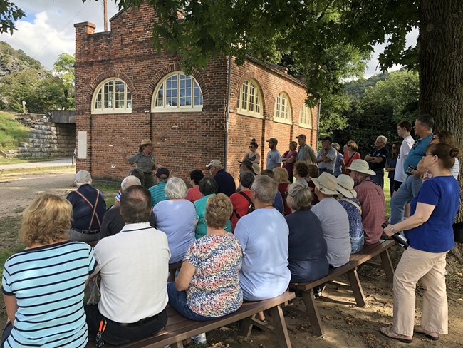 a large group of people seated and standing near the John Brown Fort brick building, while a ranger speaks to them